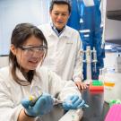 A young female scientist in a lab coat and goggles pipettes a sample while a male scientist observes in a well-equipped laboratory.
