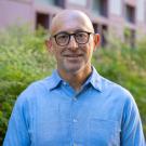 Person smiling outdoors in a light blue collared shirt, wearing glasses, greenery and building in background.