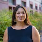 Person smiling outdoors in a sleeveless black top with long brown hair, greenery and building behind.