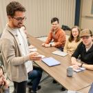 Nate Herrera, a UC Davis graduate student, smiles and gestures while leading a small group discussion in a classroom. Seated students listen attentively, some taking notes, with laptops and notebooks on the table.