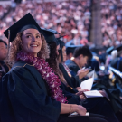 A smiling graduate wearing a flower lei sits among fellow graduates in a large venue, surrounded by a cheering audience.