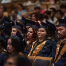 Graduates in blue and gold caps and gowns sit attentively in an auditorium, with one student's cap featuring a glowing red light.