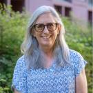Person smiling outdoors wearing a blue patterned top, long gray hair and glasses, greenery behind.