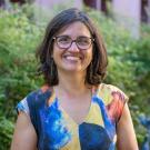 Person smiling outdoors in a colorful patterned top, glasses, and short dark hair, greenery behind.