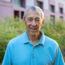 Person smiling outdoors in a light blue polo shirt, gray hair, greenery and building background.