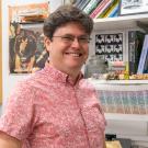 A smiling man wearing glasses and a red patterned shirt stands in a colorful office or lab space. Behind him are shelves filled with books, framed artwork, and quirky items.