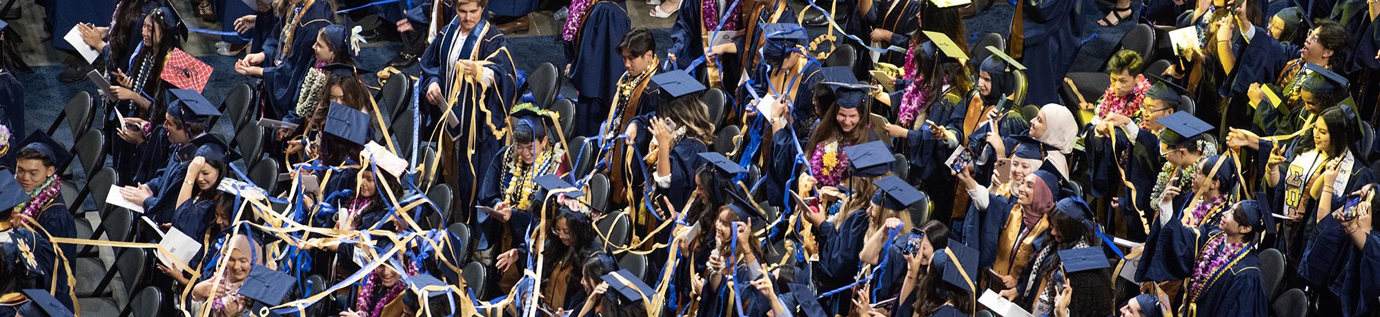 UC Davis graduates in navy blue and gold regalia celebrate at commencement, tossing caps and cheering as blue and gold streamers fill the air.