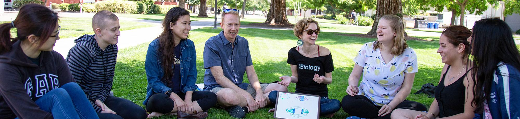  A group of students and two instructors sit in a circle on a grassy lawn, engaged in conversation and smiling. One instructor wears a black “iBiology” shirt and gestures while speaking. A small whiteboard with colorful diagrams rests on the ground in front of the group. Trees and campus buildings are visible in the background.